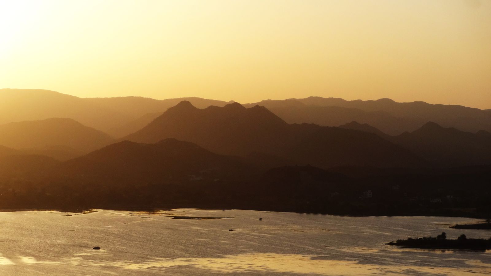 The sun setting over Lake Pichola in Udaipur, with boats on the water and hills beyond